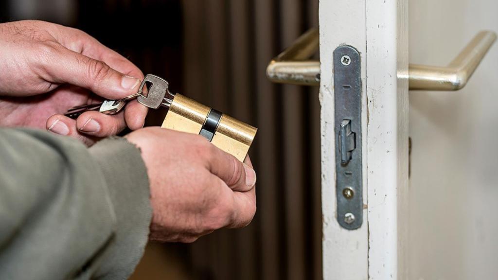 A person holds a brass-colored cylinder lock with a key inserted, positioned next to a white door frame with a metal handle and a locking mechanism.